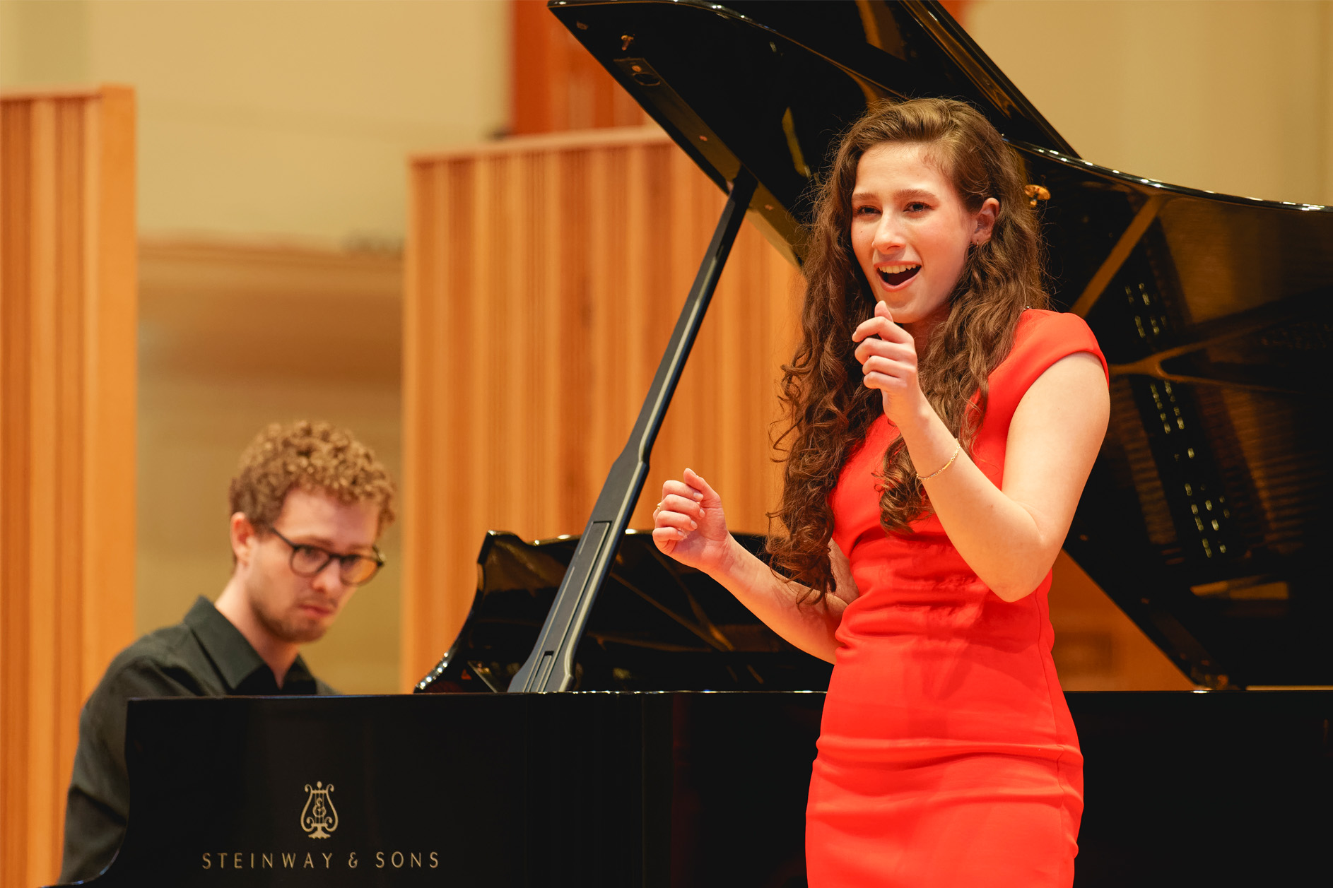 A women wearing a red dress, singing on stage, with a wooden panelled background, with a piano accompanist.